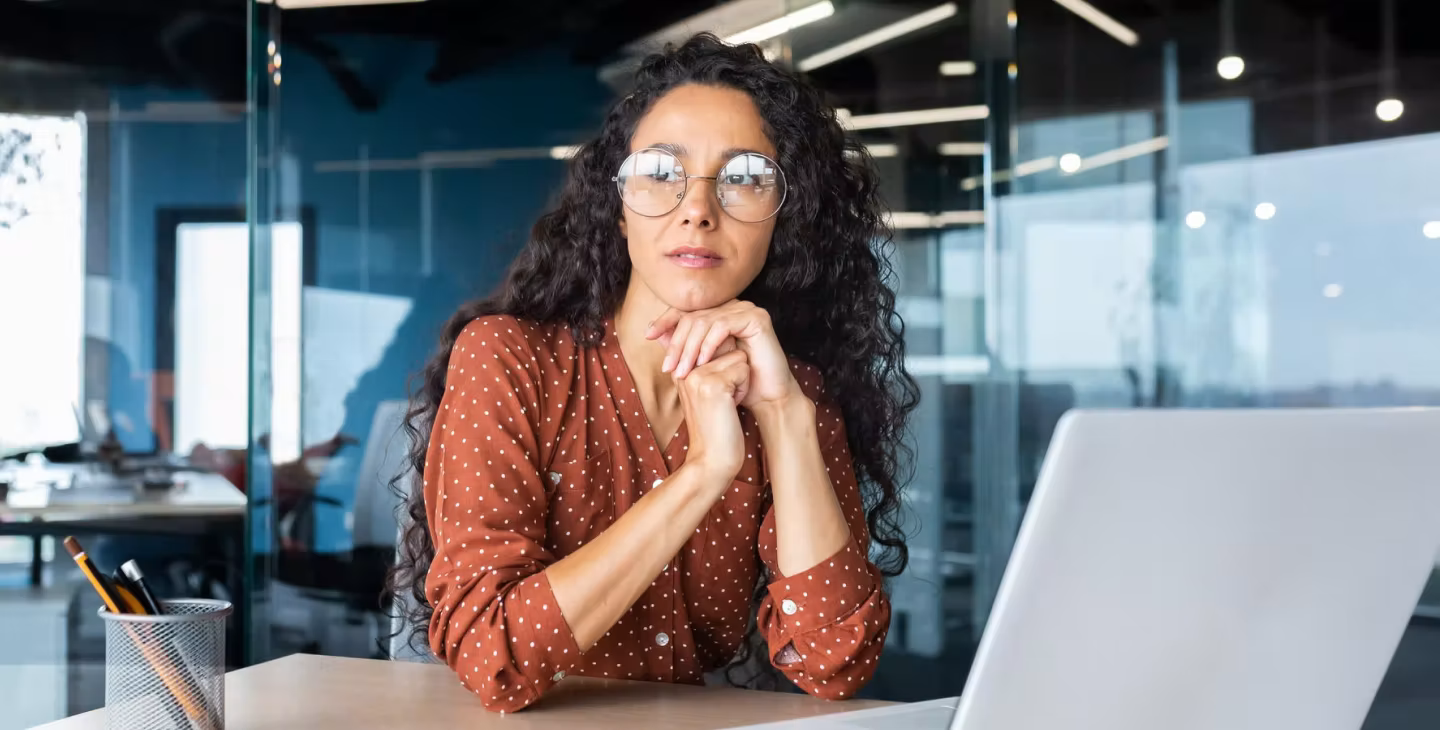 Femme avec des lunettes rondes, assise à son bureau devant un ordinateur portable, regardant pensivement au loin dans un environnement de bureau moderne.